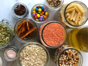 Several mason jars of various sizes, shot from above, each containing bulk food including pasta, lentils, and quinoa