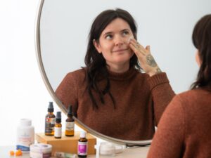 Woman At Mirror Applying Facial Moisturizer and Looking in Mirror